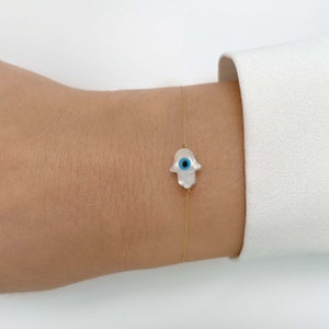 May include: A delicate gold chain bracelet with a white pearl hamsa hand charm featuring a blue evil eye.