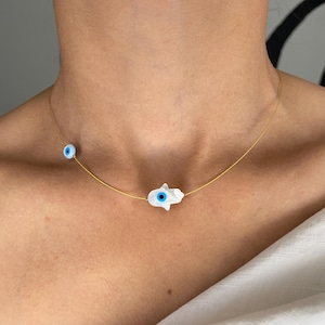 May include: A gold chain necklace with a white hamsa hand charm and a blue evil eye charm.
