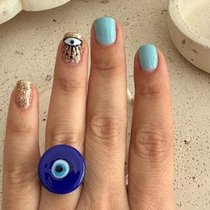 May include: A hand with light blue painted nails and a gold glitter nail. A blue eye ring is on the ring finger. The middle finger has a gold glitter nail with an eye design. The ring is a dark blue circle with a white and black eye design.