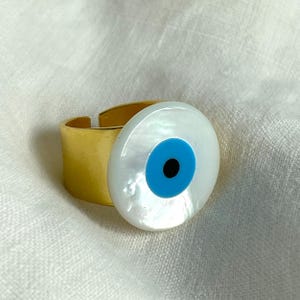 Puede incluir: Anillo dorado con un gran dije redondo con diseño de ojo turco. El dije tiene un fondo blanco nacarado, un iris azul y una pupila negra. El anillo está sobre una tela blanca.