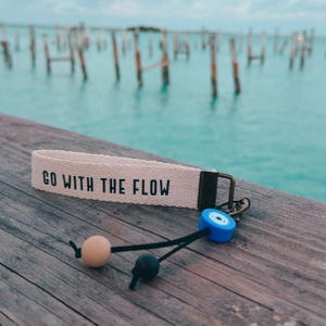 May include: A beige fabric keychain with the phrase "GO WITH THE FLOW" in black lettering. The keychain has a metal clasp and a black cord with a light beige bead, a black bead, and a blue and white bead. The background is a wooden dock and ocean.