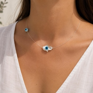 May include: A delicate necklace featuring a clear nylon cord with a central white mother-of-pearl hamsa hand charm and a small blue and white evil eye bead accent. The hamsa charm has a blue and white evil eye detail in the center of the palm.