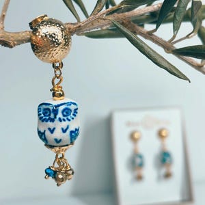 Könnte beinhalten: Goldfarbener Ohrring mit einem weißen und blauen Eulen-Design. Der eulenformige Anhänger hängt an einer goldfarbenen Kette und ist mit kleinen blauen und goldenen Perlen verziert. Der Ohrring hängt an einem Zweig.