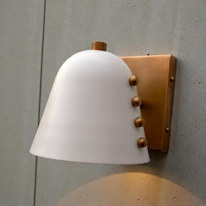 Puede incluir: Aplique de pared blanco en forma de campana con una placa trasera rectangular de color latón. El aplique tiene cuatro remaches color latón y una pequeña parte superior de madera. Montado en una pared gris.