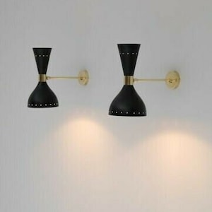 May include: Two black and gold wall sconces with a mid-century modern design. Each sconce features a black, hourglass-shaped shade with small, decorative rivets. The sconces are mounted on a wall with a gold arm and a circular gold base.