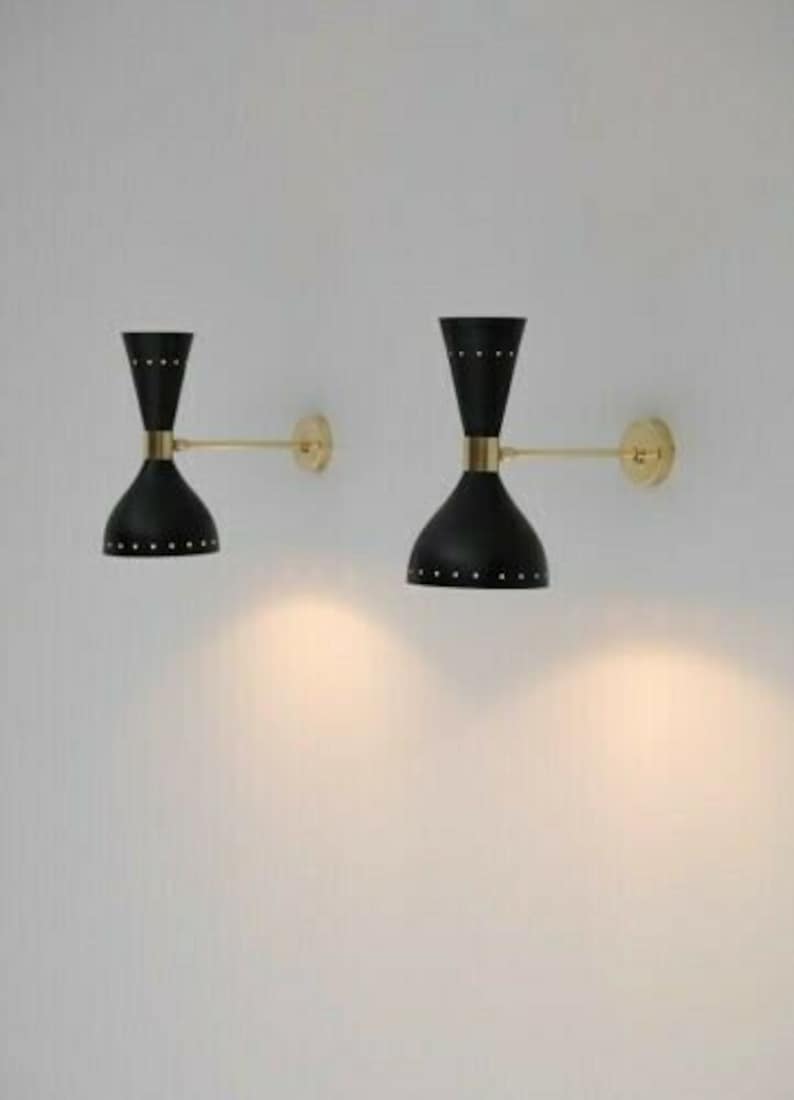 May include: Two black and gold wall sconces with a mid-century modern design. Each sconce features a black, hourglass-shaped shade with small, decorative rivets. The sconces are mounted on a wall with a gold arm and a circular gold base.