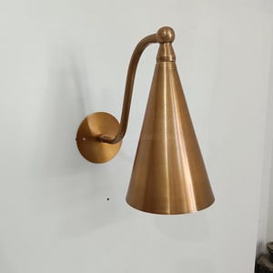 May include: A brass-colored wall sconce with a conical shade. The sconce has a curved arm and a round base. The shade is cone-shaped and tapers to a point. The sconce is mounted on a white wall.
