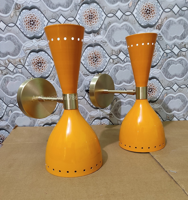 May include: Two orange mid-century modern wall sconces with brass accents. Each sconce has a conical shade with a flared top and a series of small white dots. The sconces are mounted on round brass wall plates and have a sleek, retro design.