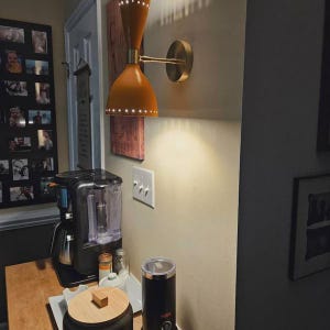 May include: A mid-century modern wall sconce with a mustard yellow shade and brass accents. The light fixture is mounted on a beige wall, illuminating the area. A coffee maker and grinder are visible on a wooden surface.