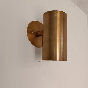 May include: A cylindrical wall sconce made of brushed brass. The sconce has a circular backplate and a straight, vertical shade. The fixture is mounted on a white wall. The text "antiquegiftercrafts786" is visible on the backplate.
