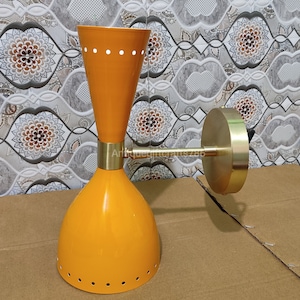 May include: An orange wall sconce with a conical shade and a cylindrical top. The sconce has a brass arm and mounting plate. The shade has small circular cutouts near the top and bottom. The sconce is a mid-century modern design.