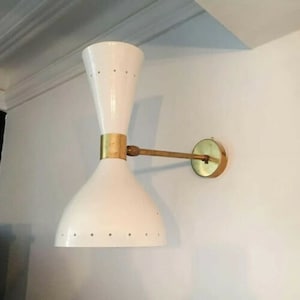 May include: A white wall sconce with a brass arm and accents. The sconce has a double-cone design with small, evenly spaced holes near the bottom. The fixture is mounted on a white wall.