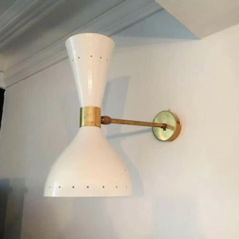 May include: A white wall sconce with a brass arm and accents. The sconce has a double-cone design with small, evenly spaced holes near the bottom. The fixture is mounted on a white wall.