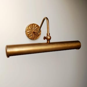 May include: A brass-colored picture light fixture. The fixture has a round wall mount with a floral design, a curved arm, and a cylindrical light bar. The light is designed to illuminate artwork or photographs.
