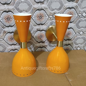 May include: Two orange wall sconces with a mid-century modern design. Each sconce has a conical shade with a white interior and a brass-colored mounting plate. The sconces have a series of small holes and white dots for added detail. The text "antiquegiftcrafts786" is visible.