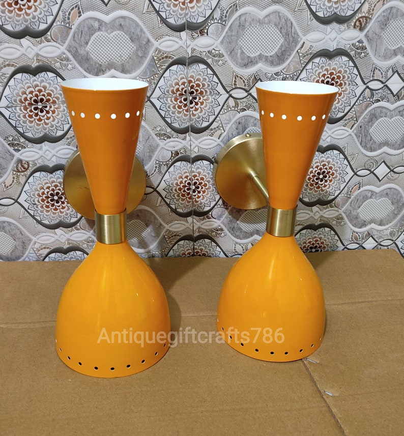 May include: Two orange wall sconces with a mid-century modern design. Each sconce has a conical shade with a white interior and a brass-colored mounting plate. The sconces have a series of small holes and white dots for added detail. The text "antiquegiftcrafts786" is visible.
