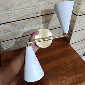 Puede incluir: Aplique de pared moderno con acabado en latón y dos pantallas blancas en forma de cono. El aplique tiene una base circular y un brazo giratorio, que ofrece iluminación ajustable. El diseño es elegante y minimalista.