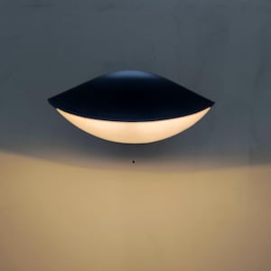 Puede incluir: Una moderna lámpara de pared negra con un diseño curvo hacia abajo. La lámpara tiene un acabado liso y mate y emite una luz suave y cálida desde su borde inferior. Está montada en una pared de color neutro.
