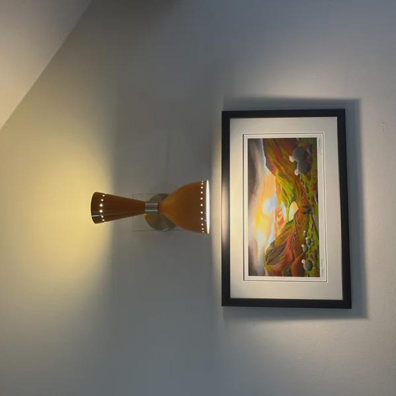 May include: A mid-century modern wall sconce with a brown and tan finish, mounted on a silver base. The sconce has a double-cone design with small perforations for light. A framed artwork hangs nearby.