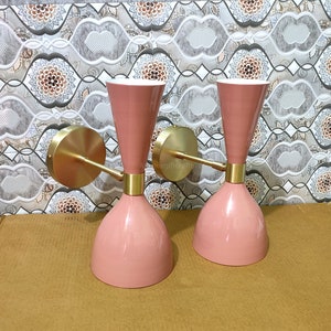 May include: Two pink wall sconces with brass accents. The sconces have a conical base, a cylindrical middle section, and a flared top. The brass arms extend from a circular base. The sconces are mounted on a patterned wall.