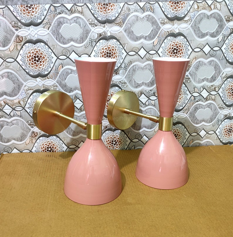 May include: Two pink wall sconces with brass accents. The sconces have a conical base, a cylindrical middle section, and a flared top. The brass arms extend from a circular base. The sconces are mounted on a patterned wall.