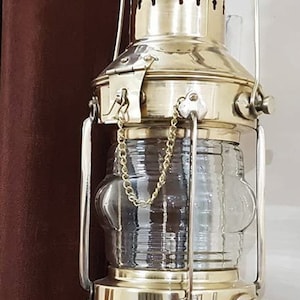 Könnte beinhalten: Eine Messing-Schiffslaterne mit einer klaren Glasscheibe und einer Kette, die von oben hängt. Die Laterne ist an einer Metallhalterung befestigt.