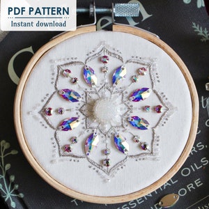 The title page of the Crystal Snowflake shows the finished embroidery centered on a black book. A vile of white seed beads sits to the right of the hoop, and DMC Diamant thread sits in top left corner under the PDF Pattern/Instant download sticker.