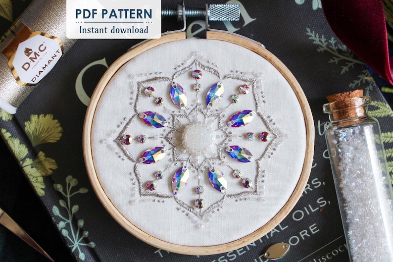 The title page of the Crystal Snowflake shows the finished embroidery centered on a black book. A vile of white seed beads sits to the right of the hoop, and DMC Diamant thread sits in top left corner under the PDF Pattern/Instant download sticker.