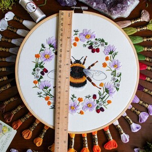 The Bumble Bee hoop on dark wood with a ruler for scale shows the hoop is 9 inches in diameter. The DMC thread used in this hoop is behind the hoop fanning outward. Flowers, moss, gems, and tarot cards are used as decor on the edge of the photo.