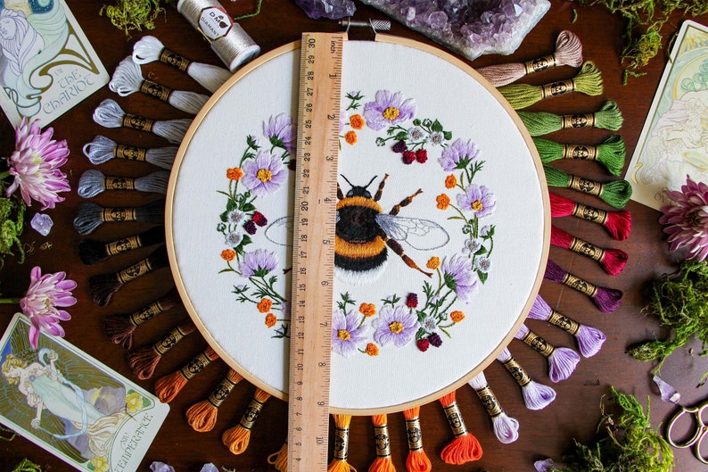 The Bumble Bee hoop on dark wood with a ruler for scale shows the hoop is 9 inches in diameter. The DMC thread used in this hoop is behind the hoop fanning outward. Flowers, moss, gems, and tarot cards are used as decor on the edge of the photo.