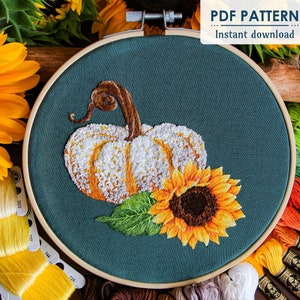 May include: Embroidery pattern for a pumpkin and sunflower. The pattern is on a teal background and includes a white pumpkin with a brown stem and a yellow sunflower with a brown center. The pattern is in a hoop and there are embroidery floss spools in the background.