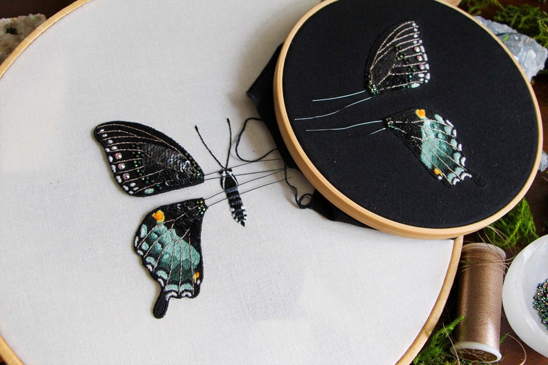 Closer view of 2 embroidery hoops of the 3D butterfly before assembly. One hoop with the butterfly wings on black fabric, and a larger hoop with the butterfly body on white fabric and cut out left side wings.