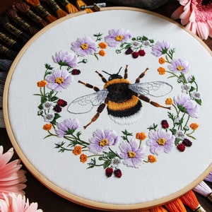 A 3/4 view of the Bumble Bee hoop with DMC thread colors radiating outward from underneath the hoop. The hoop is on a dark wood table and has pink Gerber daisy flowers and amethyst gems in the background for decoration.