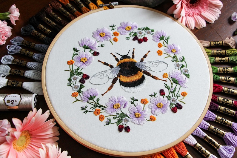 A 3/4 view of the Bumble Bee hoop with DMC thread colors radiating outward from underneath the hoop. The hoop is on a dark wood table and has pink Gerber daisy flowers and amethyst gems in the background for decoration.