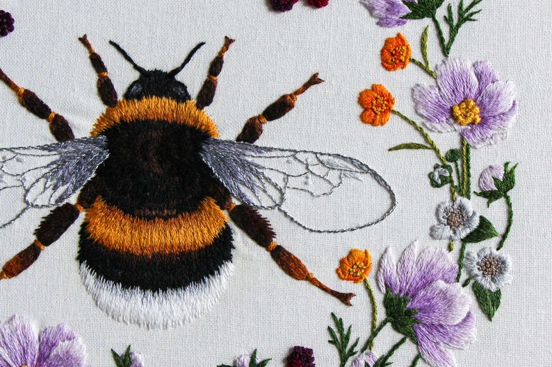 A close up of the embroidered bumble bee and flowers from the right side of the hoop uses thread painting techniques to embroider this design. Includes purple cosmos, grey blackberry flowers, and orange buttercups on a white fabric ground.