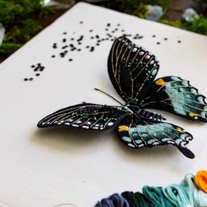 Three quarter view of the 3D butterfly mounted onto the frame backing, showing the wings are 3D. Styled with moss, gemstones, DMC thread, and embroidery materials used in this pattern.