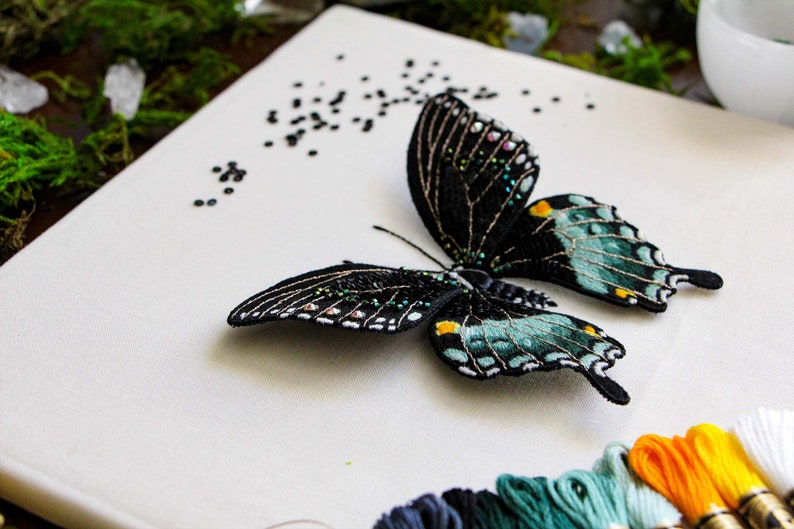 Three quarter view of the 3D butterfly mounted onto the frame backing, showing the wings are 3D. Styled with moss, gemstones, DMC thread, and embroidery materials used in this pattern.