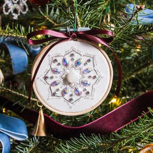 K&ouml;nnte beinhalten: Ein wei&szlig;er Stoff-Weihnachtsbaumschmuck mit einem Schneeflockenmuster, das mit schillernden Perlen und Pailletten bestickt ist. Der Schmuck h&auml;ngt an einem Weihnachtsbaum mit rotem und blauem Band.