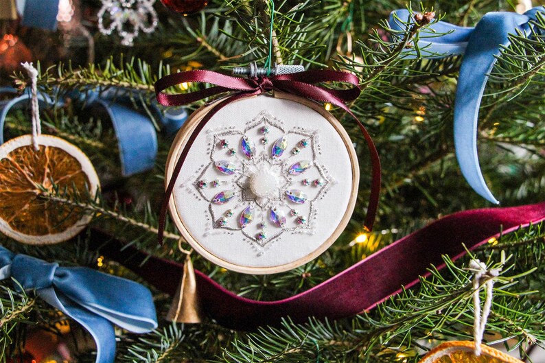 K&ouml;nnte beinhalten: Ein wei&szlig;er Stoff-Weihnachtsbaumschmuck mit einem Schneeflockenmuster, das mit schillernden Perlen und Pailletten bestickt ist. Der Schmuck h&auml;ngt an einem Weihnachtsbaum mit rotem und blauem Band.