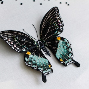 Three quarter view of the finished 3D butterfly made from thread painting embroidery and beading, on a white fabric background. Shows the wings are 3D above the white fabric.