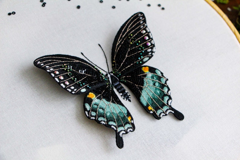 Three quarter view of the finished 3D butterfly made from thread painting embroidery and beading, on a white fabric background. Shows the wings are 3D above the white fabric.