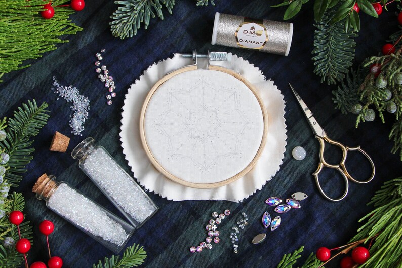 The hoop before stitching, centered in the photo, has the pattern lightly traced in pencil. The materials, beads, and thread used in the pattern surround the hoop. And Christmas greenery create a frame along the edges.
