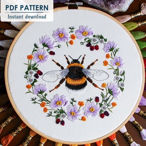 The Bumble Bee hoop art is centered in the photo, showing the thread painted bumble bee surrounded by a wreath of cosmos, buttercups, and blackberry flowers. The DMC thread used in this hoop is behind the hoop fanning outward. This is a pdf pattern.