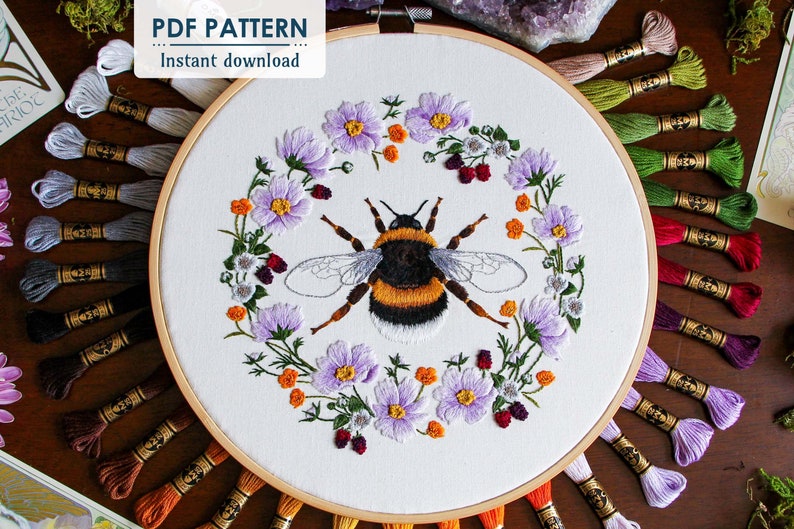 The Bumble Bee hoop art is centered in the photo, showing the thread painted bumble bee surrounded by a wreath of cosmos, buttercups, and blackberry flowers. The DMC thread used in this hoop is behind the hoop fanning outward. This is a pdf pattern.