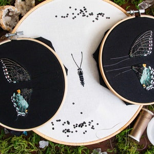 3 embroidery hoops of the 3D butterfly before assembly. 2 hoops with the butterfly wings on black fabric and a larger hoop with the butterfly body on white fabric. Styled with moss and embroidery materials