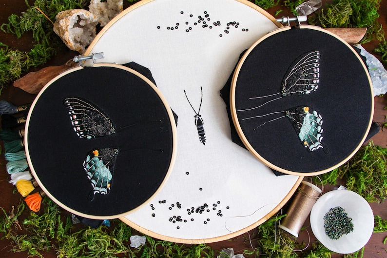 3 embroidery hoops of the 3D butterfly before assembly. 2 hoops with the butterfly wings on black fabric and a larger hoop with the butterfly body on white fabric. Styled with moss and embroidery materials