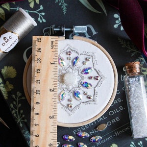 Measuring the finished Crystal Snowflake in it’s 3.5 inch hoop. The finished embroidery sits centered on a black book. A vile of white seed beads sits to the right of the hoop, and DMC’s silver Diamant thread sits in the top left corner.