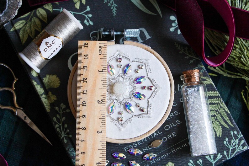 Measuring the finished Crystal Snowflake in it’s 3.5 inch hoop. The finished embroidery sits centered on a black book. A vile of white seed beads sits to the right of the hoop, and DMC’s silver Diamant thread sits in the top left corner.