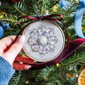 K&ouml;nnte beinhalten: Ein wei&szlig;er Stoff-Weihnachtsbaumschmuck mit einem Schneeflockenmuster, das mit schimmernden Pailletten bestickt ist. Der Schmuck h&auml;ngt an einem Weihnachtsbaum mit anderem Schmuck.