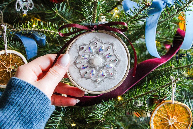 K&ouml;nnte beinhalten: Ein wei&szlig;er Stoff-Weihnachtsbaumschmuck mit einem Schneeflockenmuster, das mit schimmernden Pailletten bestickt ist. Der Schmuck h&auml;ngt an einem Weihnachtsbaum mit anderem Schmuck.